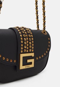 Mustan nahkalaukun, jossa on kultaisia niittejä ja suuri kultainen "G"-logo. Mukana on kierretty kultaketju sekä tekstuuritettu yksityiskohta.