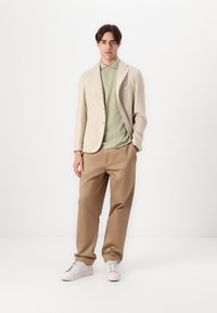 Blazer beige clair en tissu texturé, porté sur un polo vert doux. Un pantalon tan ample et des baskets blanches complètent le look.