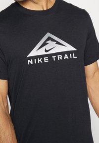 Camisa de trail negra de Nike con un logo triangular blanco que presenta una montaña estilizada y el swoosh de Nike en la parte superior del torso de una persona.