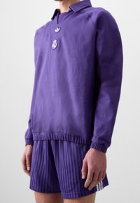 Lila sporttröja med krage, som har en broderad emblem och matchande randiga shorts. Mjuk tyg, avslappnad passform, elastiska mudd.