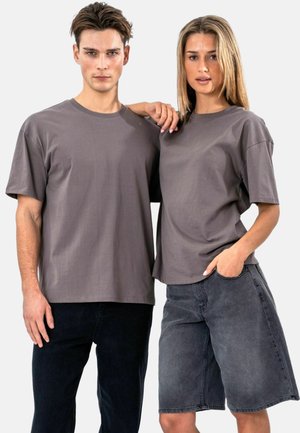 Młody mężczyzna i kobieta stoją obok siebie, ubrani w proste szare oversize'owe koszulki; kobieta ma rękę w kieszeni, a jej ramię spoczywa na ramieniu mężczyzny.