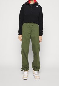Pullover in pile nero con mezzo zip, pantaloni cargo verdi con tasche laterali e sneakers color sabbia con lacci bianchi. Materiali morbidi e vestibilità rilassata.