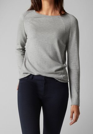 Vrouw draagt een nauwsluitende lichtgrijze trui met lange mouwen, ingestopt in donkerblauwe, smalle broek, tegen een effen achtergrond.