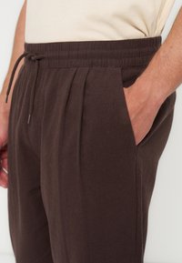 Homme portant un pantalon marron foncé avec une taille élastique et un cordon, main placée dans la poche avant droite.