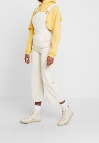 Salopette in corduroy bianco con spalline regolabili, abbinata a una felpa gialla e a sneaker alta color crema. Taglio dritto e ampio.
