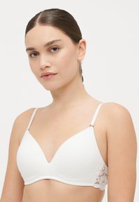 Soutien-gorge blanc avec une coque lisse et moulée et un décolleté en V profond. Des détails en dentelle sur les côtés ajoutent de la texture. Bretelles réglables en blanc.
