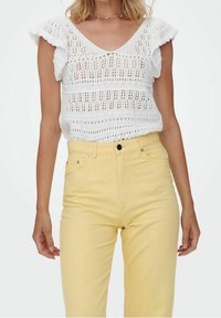 Biała dzianinowa bluzka z falbaniastymi rękawami i dekoltem w kształcie V; zestawiona z żółtymi dżinsami o wysokim stanie. Widoczne wzory teksturowe w deseniu materiału.