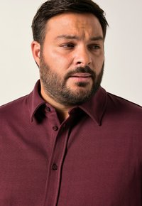 Uomo con barba che indossa una camicia borgogna con bottoni, che guarda verso sinistra su uno sfondo chiaro e uniforme.