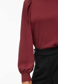 Blusa a maniche lunghe in borgogna con texture e maniche a sbuffo, abbinata a pantaloni neri a vita alta con dettaglio a bottoni.