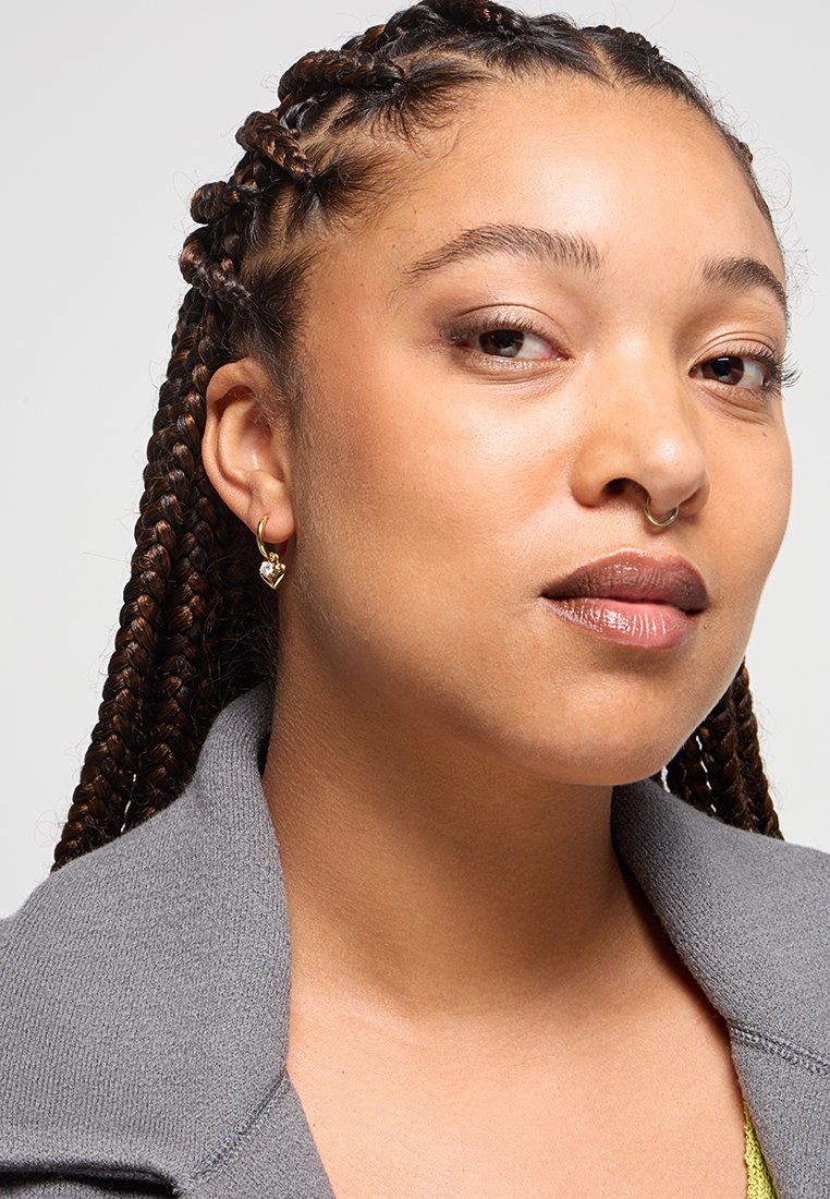 Kobieta z warkoczami, złotym sercowym kolczykiem, pierścionkiem w septum, ubrana w szary kołnierz i delikatny makijaż, pewnie patrzy w obiektyw.