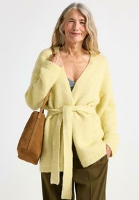 Lys gul strikket cardigan med V-hals, bælte og lange ærmer. Båret med en brun ruskinds håndtaske og en grøn top, samt løse bukser.