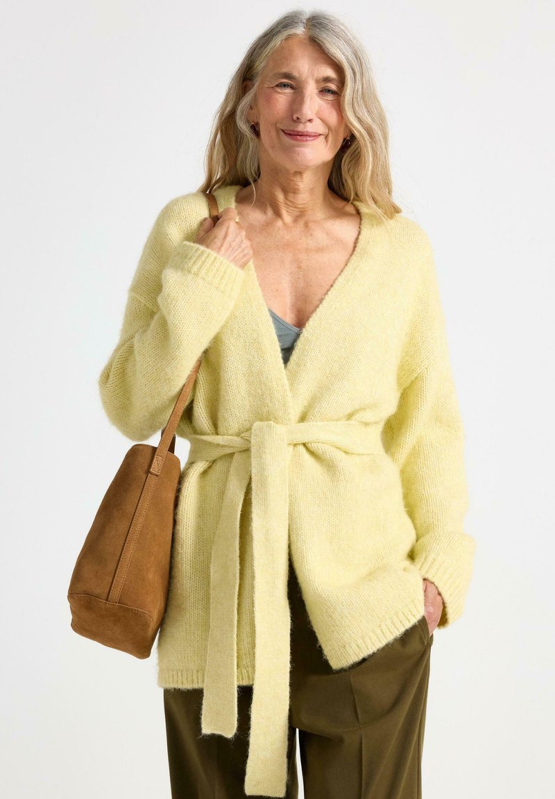 Lys gul strikket cardigan med V-hals, bælte og lange ærmer. Båret med en brun ruskinds håndtaske og en grøn top, samt løse bukser.