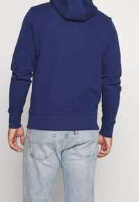 Osoba v jednoduché tmavě modré mikině a světlých džínách, stojící s výhledem zády k fotoaparátu proti jednobarevnému pozadí.