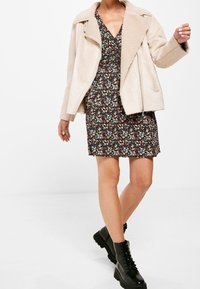 Beige nepbontjas over een bloemenjurk. De jurk heeft kleine meerkleurige bloemen op een donkere achtergrond. Zwarte veterlaarzen.