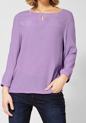 Mujer con blusa de manga larga color lila con un pequeño detalle de apertura en la parte delantera, combinada con vaqueros azul oscuro, con la mano en el bolsillo.