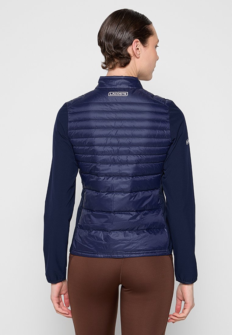 Gilet matelassé bleu marine avec une texture rembourrée, coupe slim et col haut. Présente un logo Lacoste au dos, associé à un haut à manches longues.