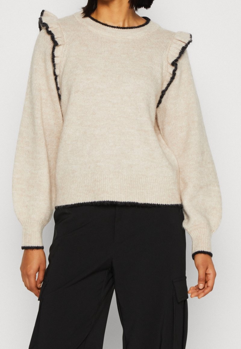 Pull beige en maille avec des détails à volants sur les épaules et un bord noir, associé à un pantalon cargo noir.