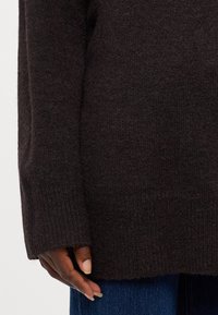 Dunkelbrauner gestrickter Pullover mit strukturiertem Finish, gerippten Bündchen und lockerer Passform. Hand auf dem unteren Saum ruhend, kombiniert mit blauen Jeans.