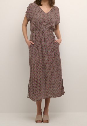 Vrouw draagt een midijurk met korte mouwen, een paisley-patroon en zijzakken, gecombineerd met beige open-toe platte schoenen.