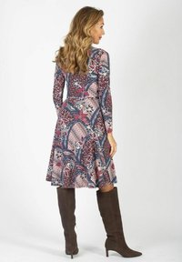 Kobieta z długimi falistymi włosami, ubrana w wzorzystą sukienkę do kolan oraz ciemno-brązowe botki na wysokim obcasie, stojąca na jednolitym tle.