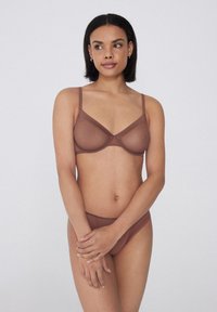 Frau mit kurzen dunklen Haaren, die einen transparenten braunen BH und passende Unterwäsche trägt, steht mit verschränkten Armen vor einem schlichten hellen Hintergrund.