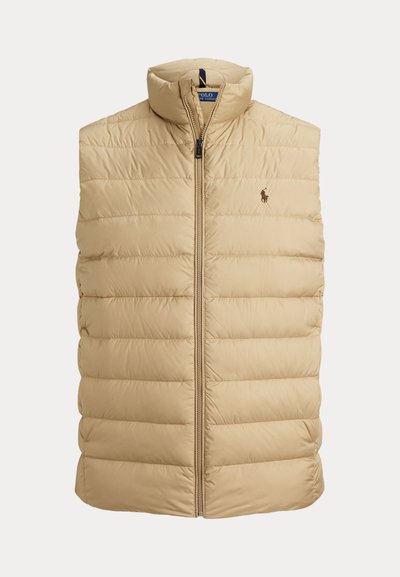 Bēša pūkaina veste ar augstu apkakli, priekšā rāvējslēdzēju un horizontālu šuvju rakstu. Uz krūtīm ir neliels izšūts logotips.