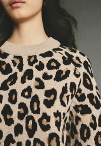 Beiger Pullover mit schwarzem und tan Leopardenmuster, ausgestattet mit einem gerippten Rundhalsausschnitt und strukturiertem Stoff. Detail an der Schulter sichtbar.