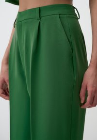 Pantalons ajustés verts en tissu lisse et texturé, avec des plis, des poches latérales et des passants de ceinture, mettant en valeur une silhouette structurée.