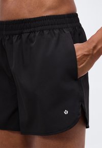 Gros plan d'une personne portant un short de sport noir avec une ceinture élastique, main dans la poche latérale, et petit logo blanc près de l'ourlet.