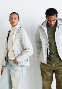 Lightweight, beige zip-up jackets with hoods, contrasted zippers, worn over a cream turtleneck and an olive-green shirt with loose denim pants.
