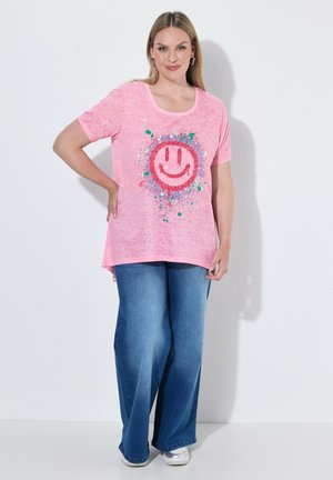Frau mit blondem Haar, die ein pinkes T-Shirt mit buntem Smiley-Design, weite blaue Jeans und weiße Schuhe trägt, steht an einer weißen Wand.