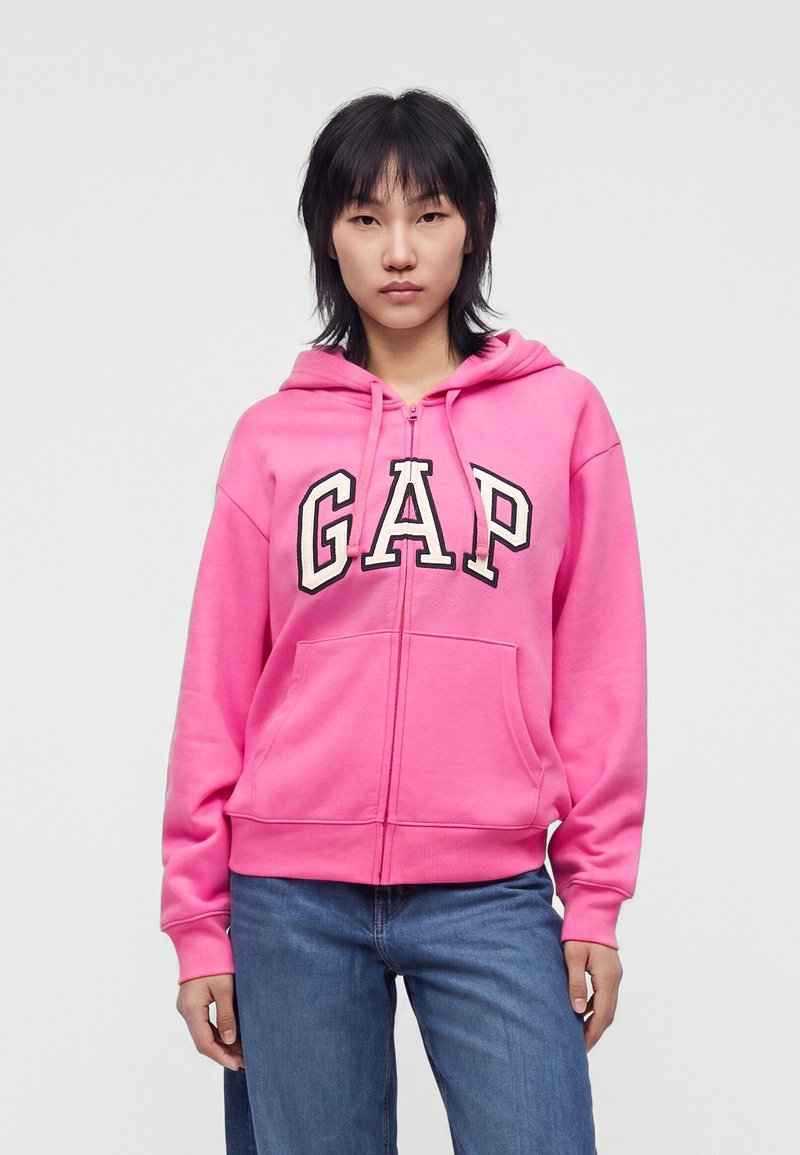 Personne portant un sweat à capuche zippé GAP rose vif et un jean bleu, debout devant un fond clair uni, face à l’avant, les mains le long du corps.