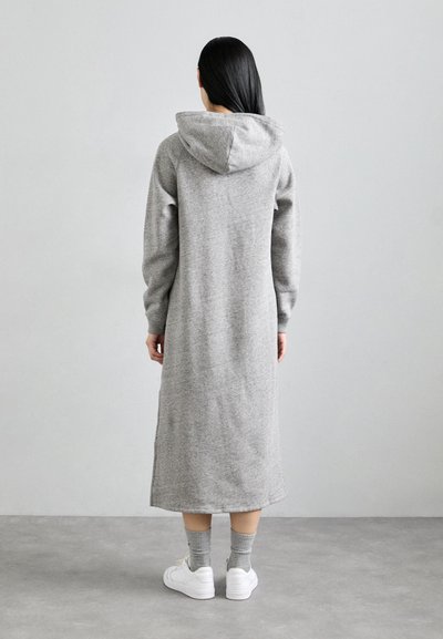Lang grijze hoodie jurk van een textuurstof, voorzien van een capuchon, lange mouwen en een rechte zoom. In combinatie met witte sneakers en grijze sokken.