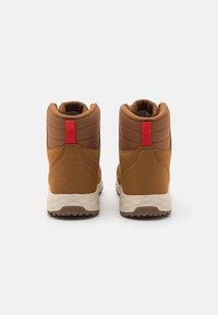 Botas de tobillo marrones con una textura suave, con acentos rojos en el tacón y una suela de goma texturizada para agarre y soporte.