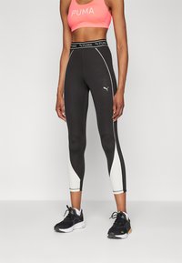 Leggings neri Puma con dettagli laterali bianchi, vita alta e cuciture riflettenti, abbinati a un crop top rosa e scarpe da ginnastica nere.