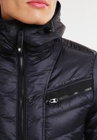 Svart pufferjacka med hög krage, quiltad textur, framställd dragkedja och en ficka med dragkedja på bröstet. Tyget är slätt och glansigt.