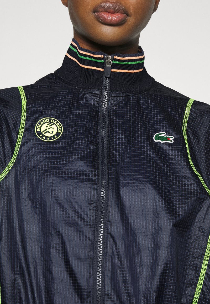 Veste zippée marine en tissu texturé, avec des accents verts et jaunes, et un logo "Roland Garros". Dotée d'un col en ribbed rayé.