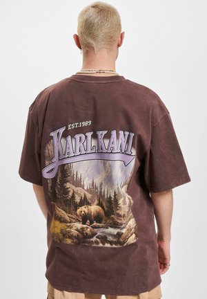 Blonde persoon die een bruin oversized T-shirt draagt met de tekst "Karl Kani" en een boslandschap met een beer bij een rivier op de achterkant.