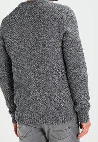 Grauer Strickpullover mit strukturiertem Muster, rundem Ausschnitt und gerippten Bündchen sowie Saum. Getragen mit grauen Hosen, was einen lässigen Stil zeigt.