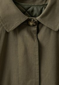 Olívazöld gombos kabát hegyes nyakkal, texturált anyagból készült és egy látható gombbal záródik. A bélésen gyémántmintázat látható.