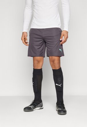 Pantalones cortos deportivos grises con detalles en morado, combinados con calcetas negras hasta la rodilla que cuentan con un logo blanco de Puma y zapatos deportivos negros.