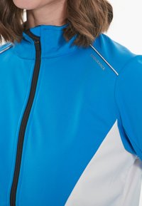 Blaue Sportjacke mit hohem Kragen, ausgestattet mit einem schwarzen Reißverschluss und weißen Akzenten. Glattes Material mit sportlichem Design. Logo auf der Schulter.