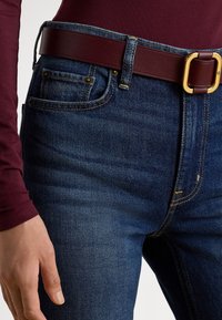 Κοντινό πλάνο ενός ατόμου που φορά μπλε τζιν τζιν, μια σκούρα κόκκινη ζώνη με χρυσή αγκράφα και σκούρο κόκκινο πουκάμισο με μακριά μανίκια, το χέρι ακουμπισμένο στο πλάι.