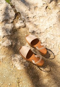 Pár béžových sandálů s otevřenou špičkou a kotníkovými pásky na drsném, skalnatém povrchu při přirozeném slunečním světle vrhajícím stíny.