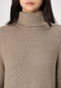 Pull beige en tricot avec un col haut et replié. Présente une surface texturée avec un côtelé vertical, créant une apparence chaleureuse.