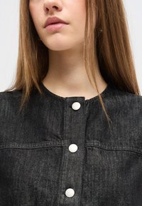 Νεαρή γυναίκα με ίσια ανοιχτό καστανά μαλλιά που φοράει μαύρο τζιν μπουφάν με τρία ορατά ασημένια κουμπιά πατιλέτας.