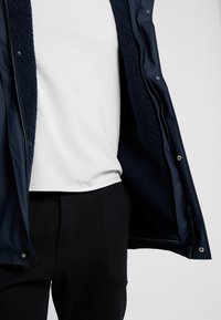 Marinejakke med fleece-for og trykknapper. Båret over en hvid t-shirt og sorte bukser. Blød tekstur tydelig indeni jakken.