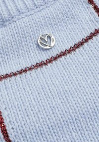 Un tissu tricoté bleu clair présente des coutures métalliques rouges et un bouton circulaire en argent avec un découpage en forme de cœur.