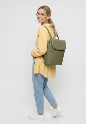 Femme souriante, vêtue d'une chemise jaune clair, d'un haut blanc, d'un jean bleu, de baskets blanches, et portant un sac à dos en cuir vert olive.