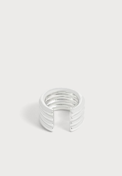 Une bague ouverte en argent poli, ornée de trois reliefs horizontaux, sur un fond clair et uni.